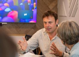 A man sitting at a desk holding his hands out in demonstration, with a woman sitting next to him watching on