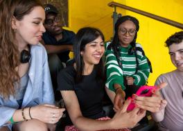 Three women and two men sitting together and looking at a mobile phone being held by one of the women