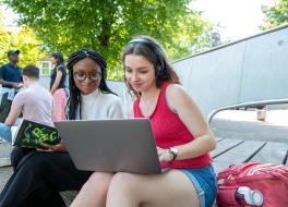 A woman in headphones showing something on her laptop to the woman sitting next to her.