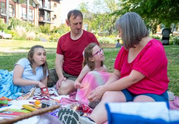 A family picnic, with a young girl speaking and her mother, father and daughter sitting listening to her