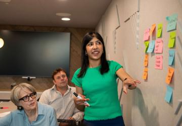 A woman standing next to a wall with post it notes on it, with a woman and a man sitting beside her, watching