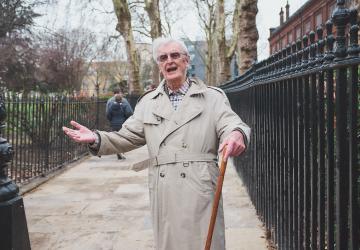 A man in a rain mac holding a walking stick holding his arms out