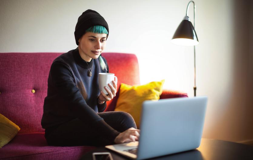 Woman on a sofa, drink in hand, typing on a laptop