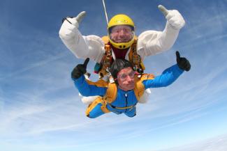 Two men doing a tandem parachute jump, both looking at the camera with their thumbs up