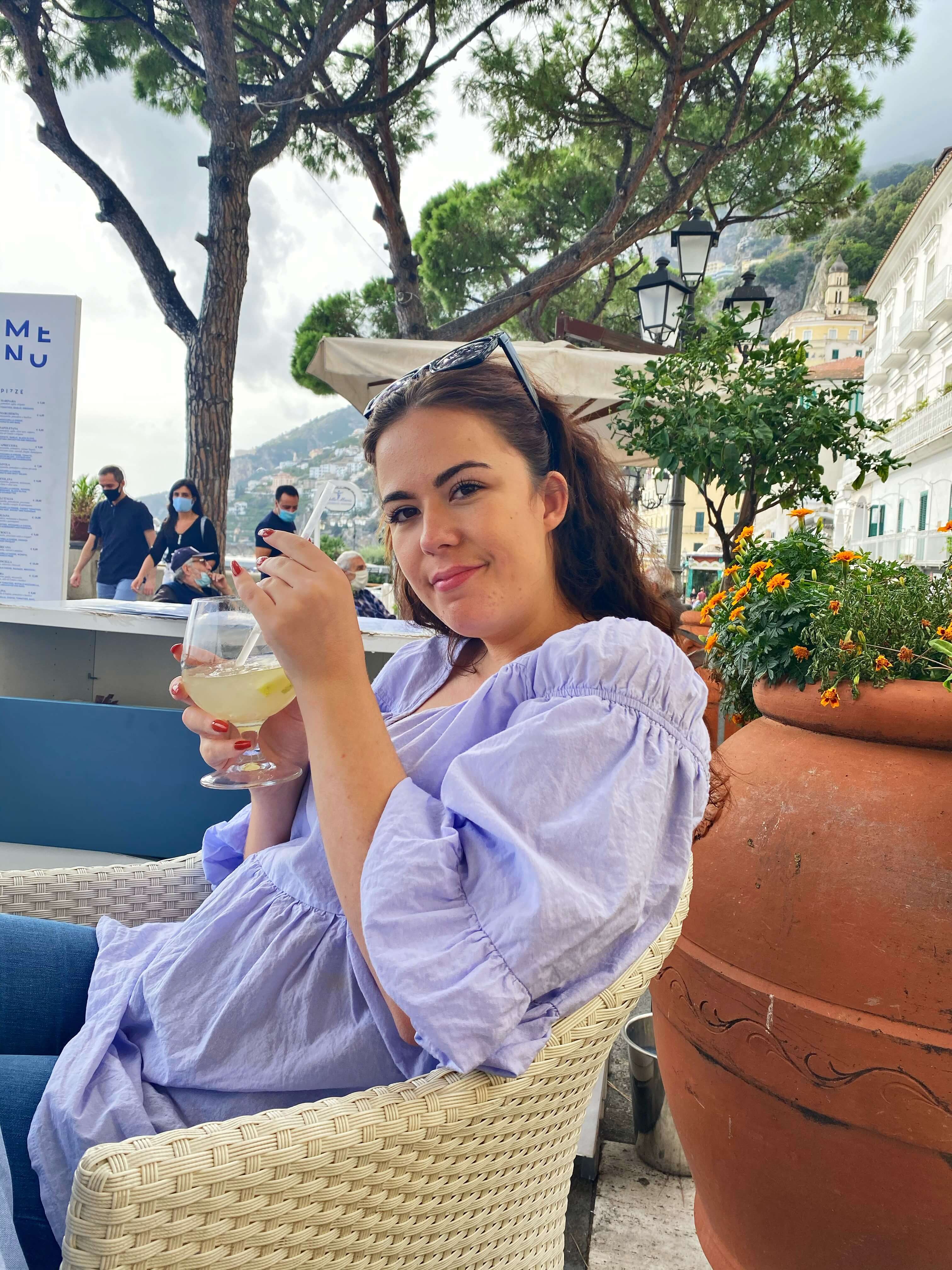 A young woman sitting in a chair in an outdoor restaurant, holding a drink